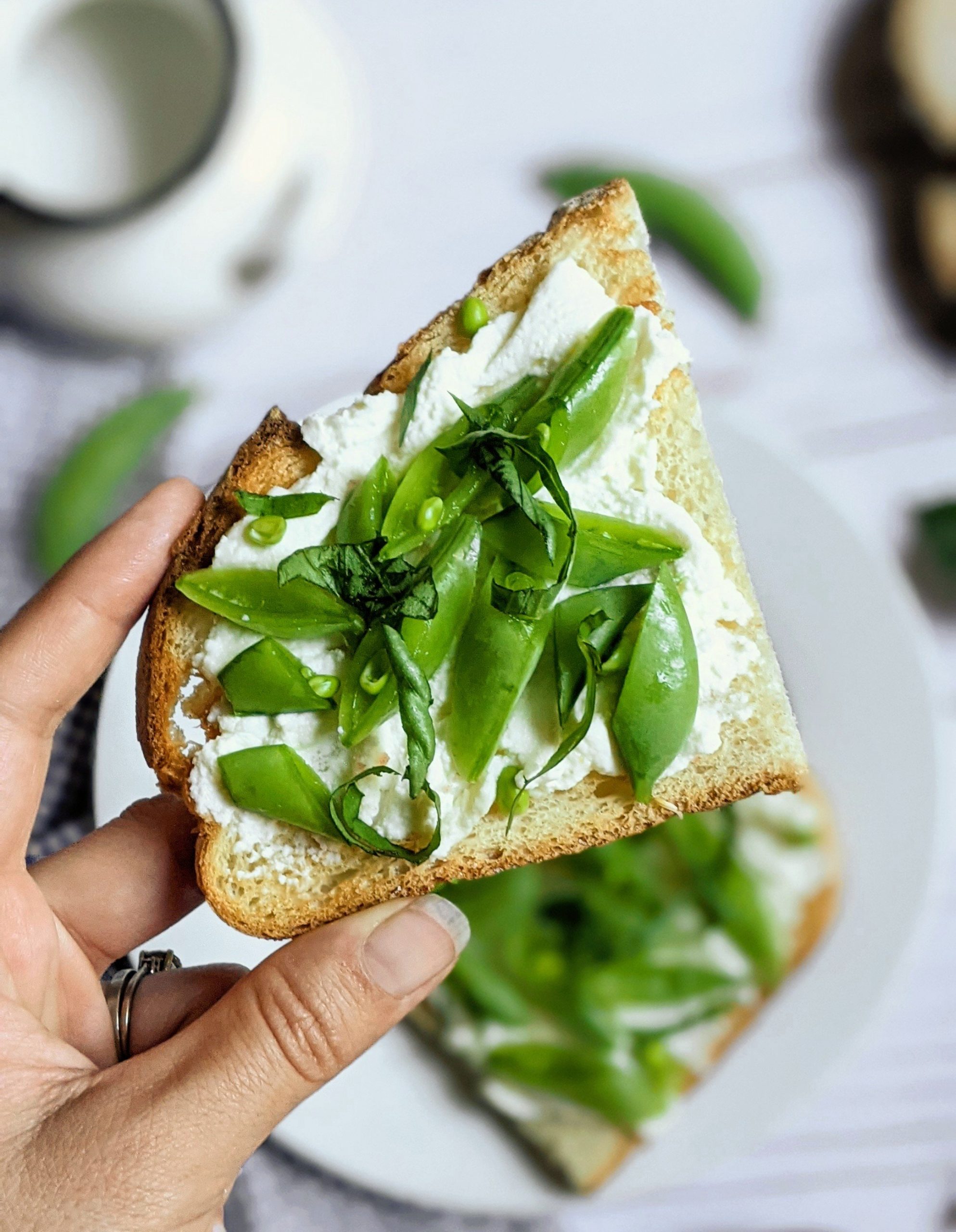 hand holding a piece of toast with snap peas ricotta and basil for breakfast with another breakfast toast on a plate