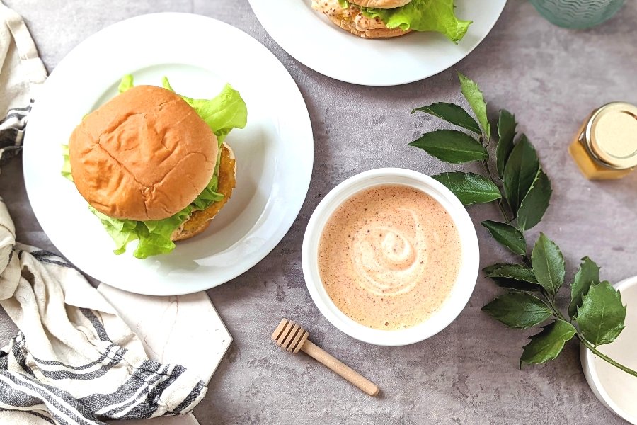 chicken sandwich sauce recipe with honey mustard mayo spices paprika and spicy chili flakes in a ramekin drizzled on a chicken sandwich with a honey dipper.