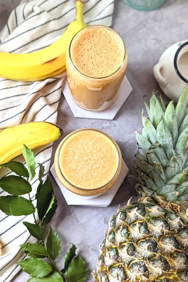 overhead shot of two vegan pineapple smoothies with banana and pineapple and leaves and greens and a striped napkin.