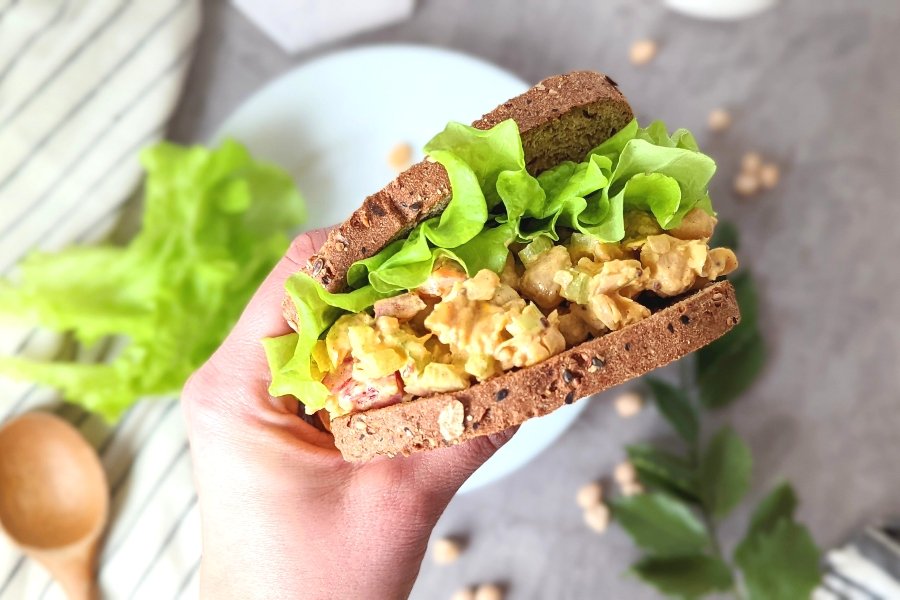 a hand holding a vegan chicken salad with garbanzo bean sandwich with the filling spilling out and curly green leaf lettuce on the sandwich with grains and oats in the bread.