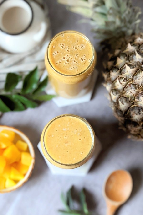 two glasses of pineapple and mango smoothie with fresh mango and a whole pineapple in a smoothie recipe for a healthy fiber rich breakfast recipe.