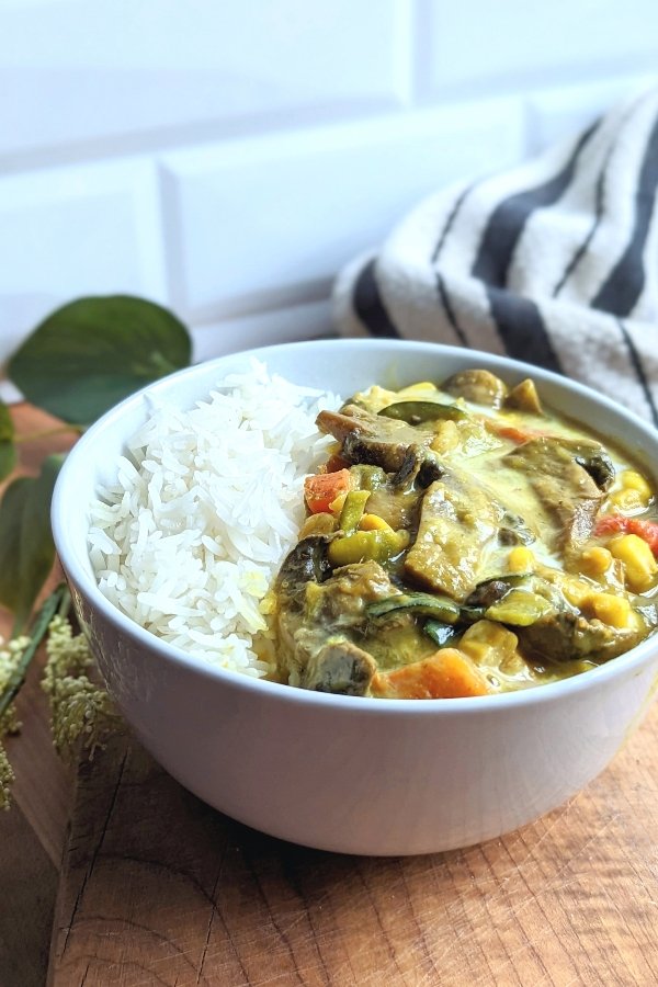 vegan curry with mushrooms in a bowl with carrots corn onion zucchini and a creamy plant based coconut milk curry sauce with fresh steamed jasmine rice and flowers in the background.