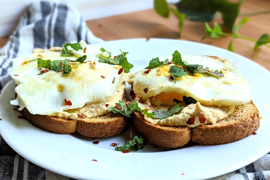 two slices of while wheat toast with hummus and fried eggs on top with fresh chopped parsley and chili flakes on a plate.