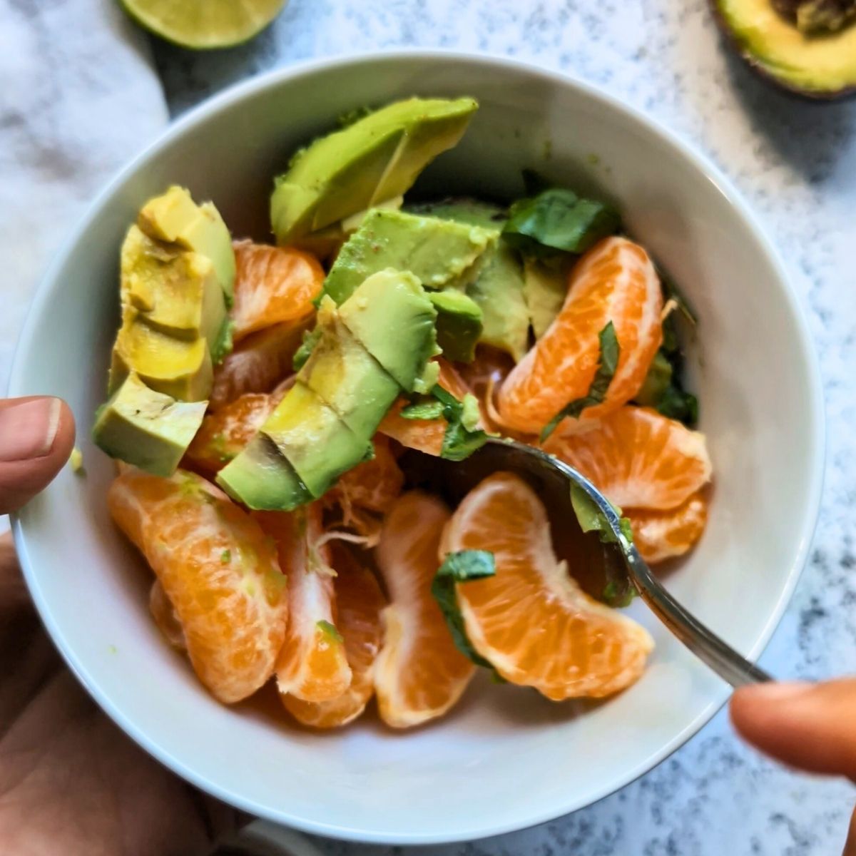 a spoon stirring a bowl of sliced orange salad with avocado and lime and fresh basil.