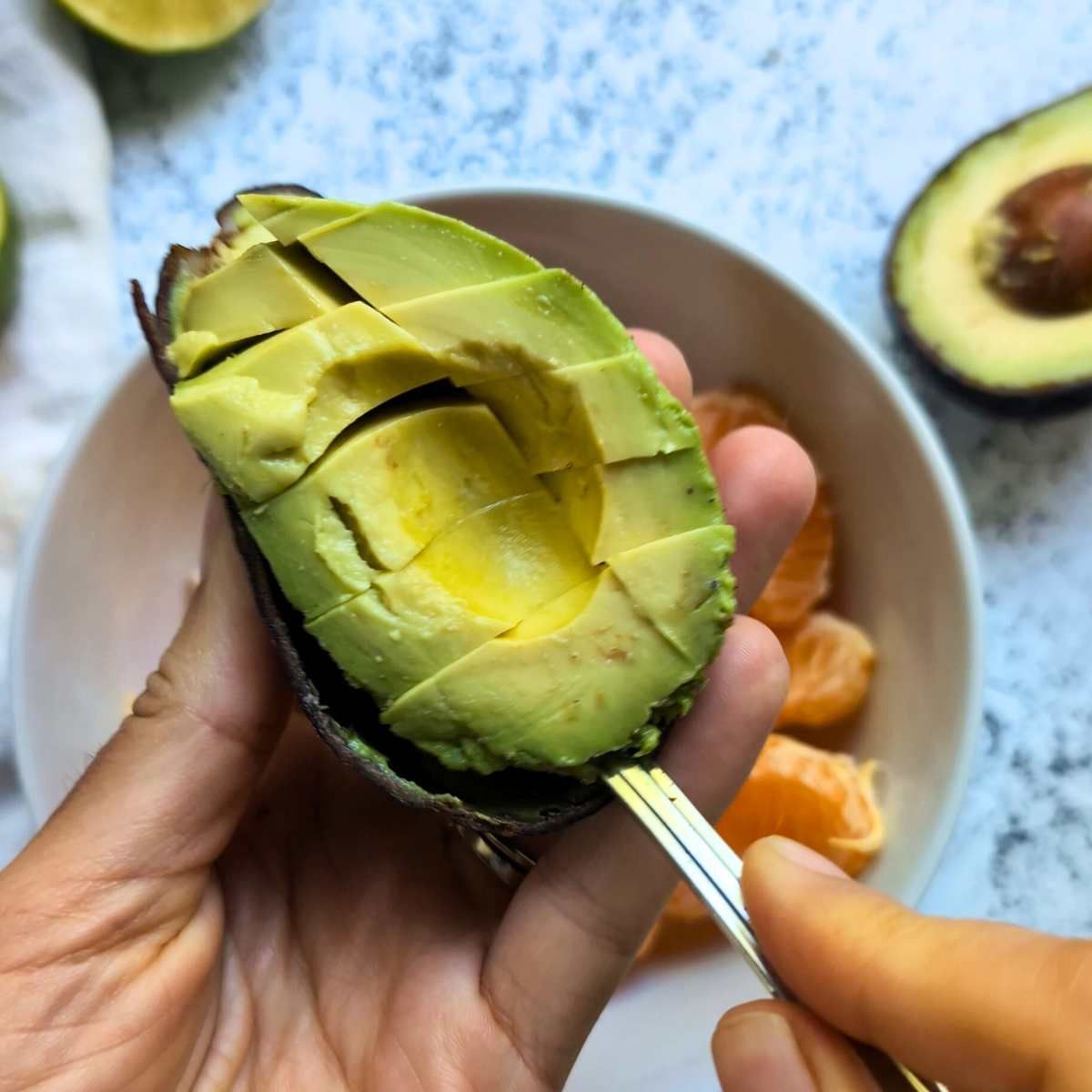 a spoon scooping out the flesh of an avocado into a bowl with orange segments.