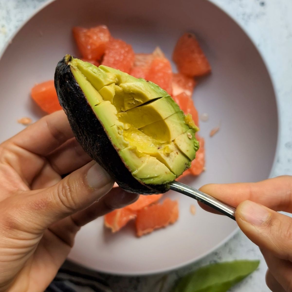 a spoon scooping out the flesh of an avocado into a bowl with grapefruit segments.