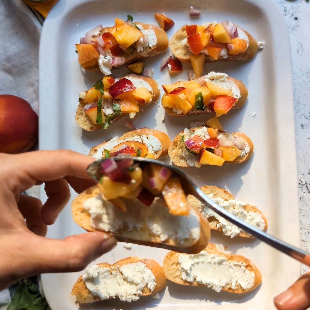 a crostini covered in goat cheese with a scoop of peach bruschetta being spooned on top.