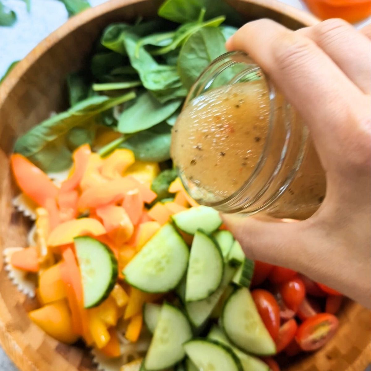 Italian dressing being poured over a large salad with chopped vegetables and farfalle pasta.