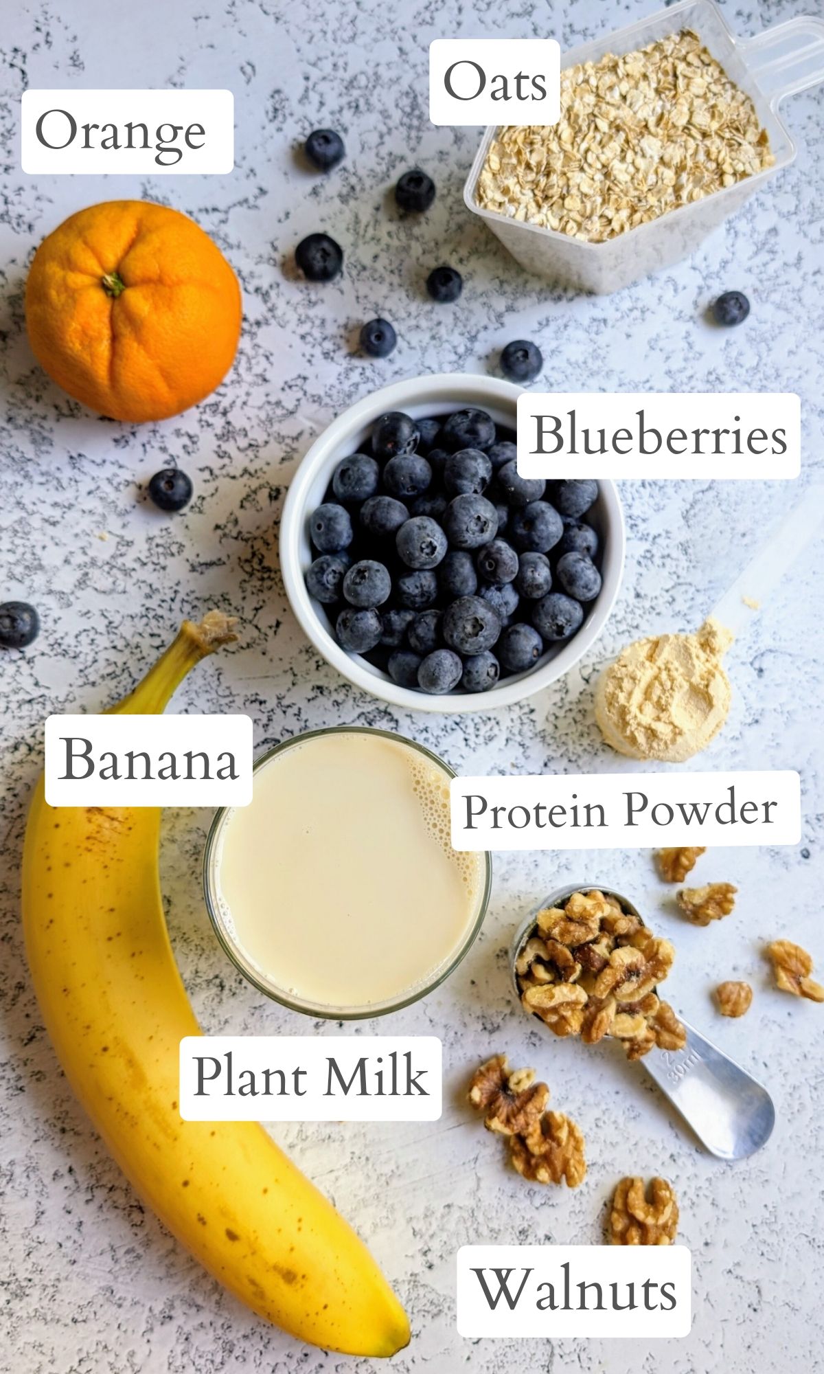 ingredients for a oatmeal blueberry smoothie laid out on a countertop: oats, an orange, vanilla protein powder, banana, plant based milk, walnuts, and fresh blueberries.