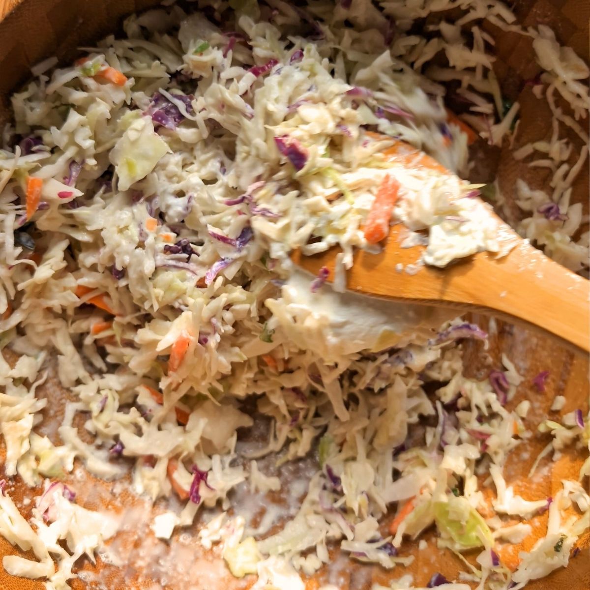 a wooden spoon stirring the sour cream into the coleslaw for a mayo free side dish.