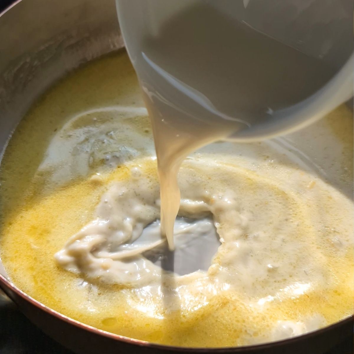 corn starch being poured into a pan of oat milk sauce with garlic and olive oil.