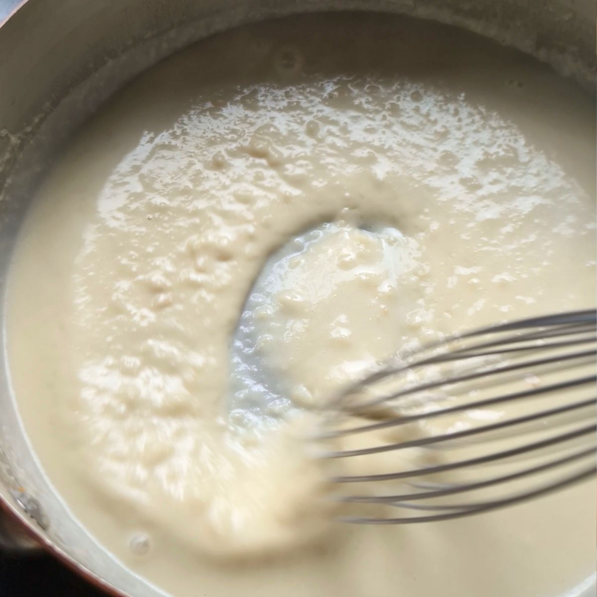 a whisk stirring a roux with milk into a thick and creamy alfredo sauce without parmesan cheese.