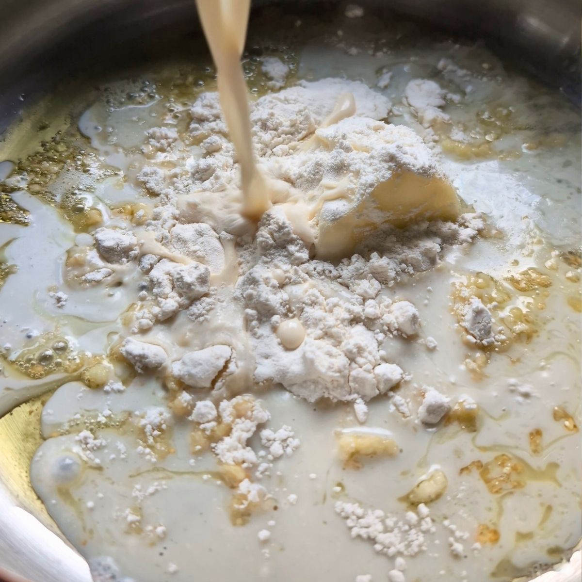 a pan with olive oil, garlic, butter, flour, and milk in a pan being stirred with a spoon.