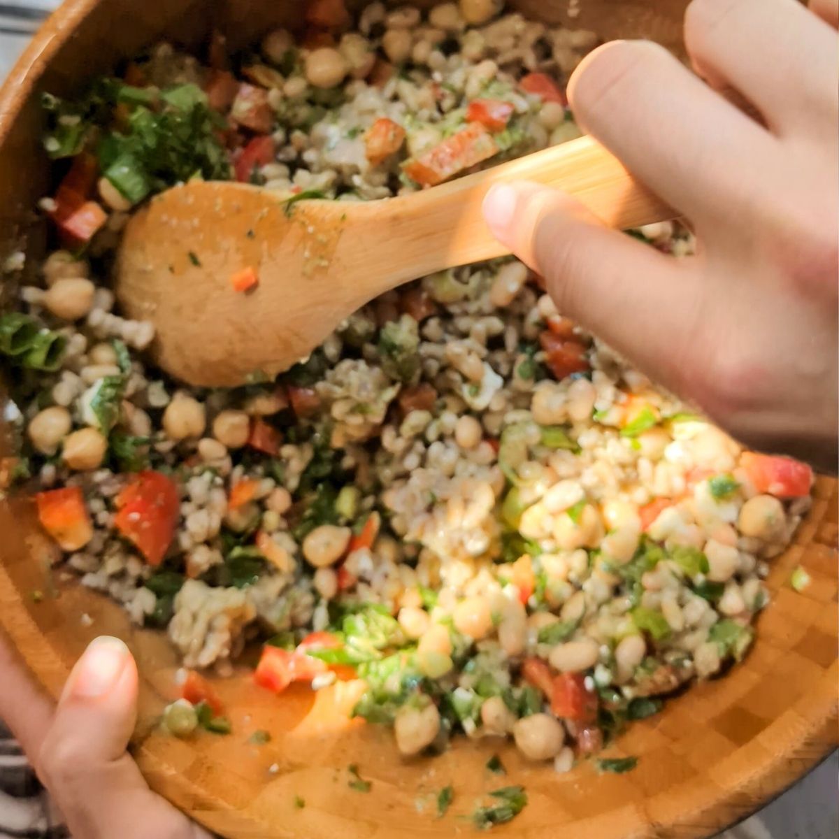 a hand and a wooden spoon mixing the bean and barley salad with the pesto basil dressing.