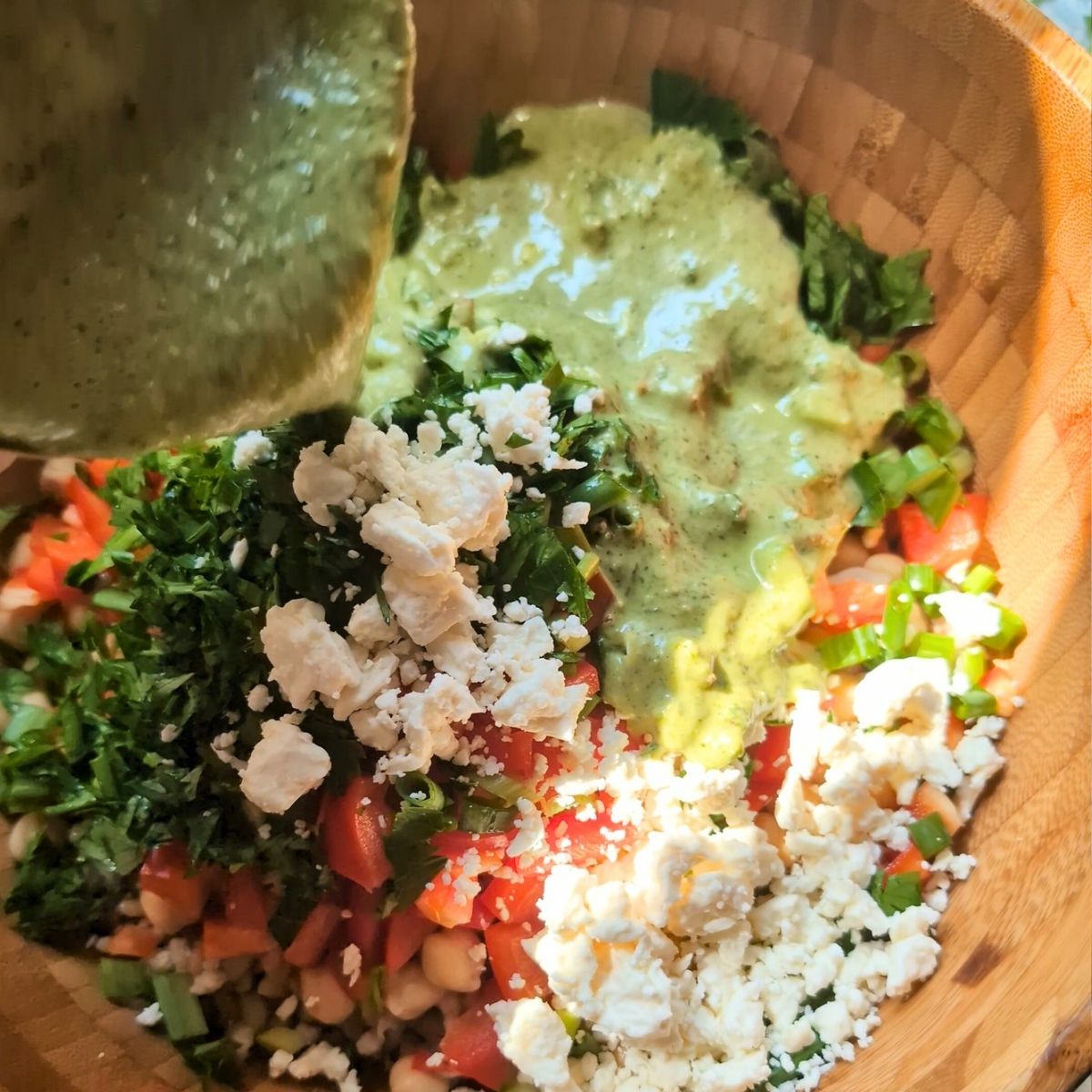 a hand pouring green pesto dressing over the bean and barley salad in a large serving bowl.