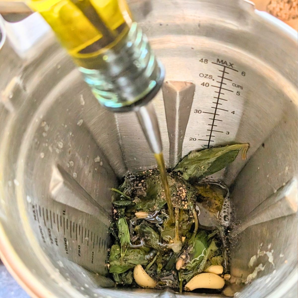a blender full of basil garlic vinegar and herbs, with olive oil being streamed in.