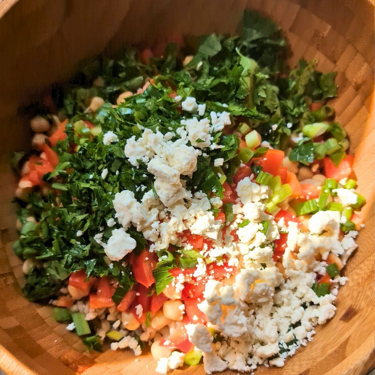 a large wooden bowl full of salad ingredients like beans, cooked barley, chopped vegetables, and feta cheese.