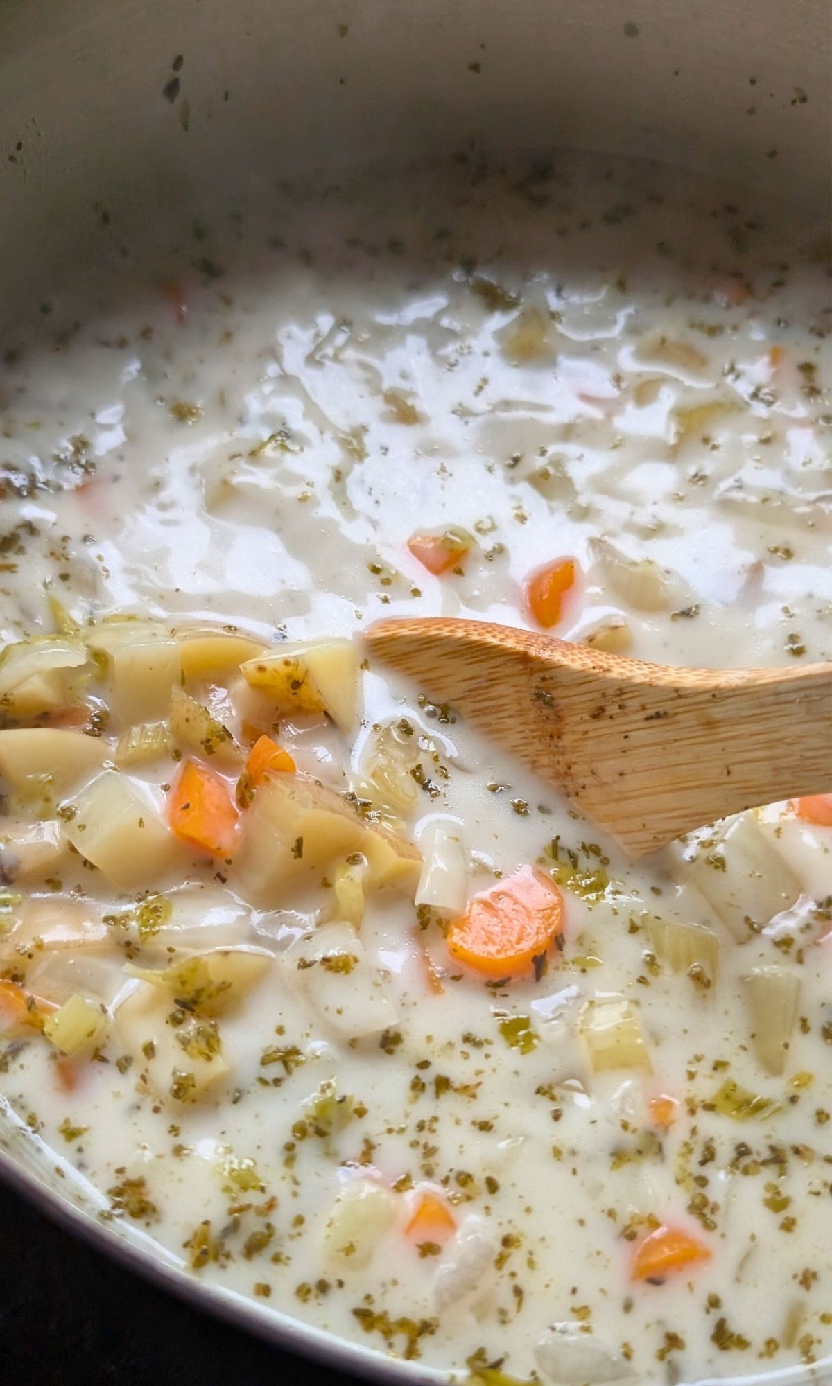 a wooden spoon stirring a homemade potato soup with coconut milk broth and fresh herbs and chunky root vegetables.