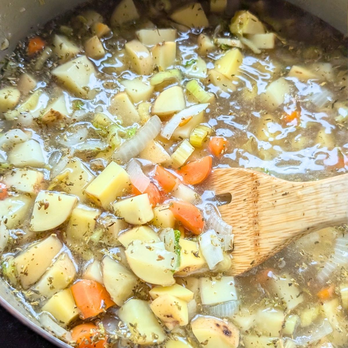 a pot of potato soup simmering in vegetable broth with chunks of yellow potatoes and carrots being stirred by a wooden spoon.