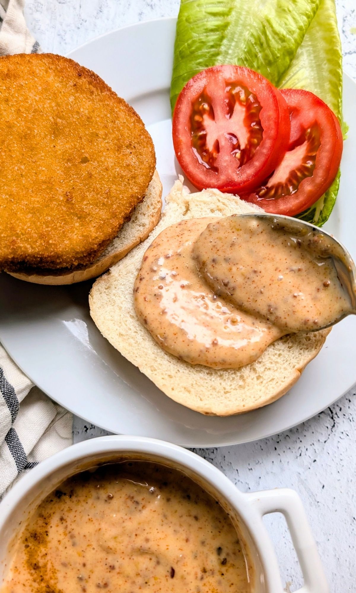 a spoon spreading chicken sandwich sauce on a bun with lettuce and tomato and a fried chicken patty.