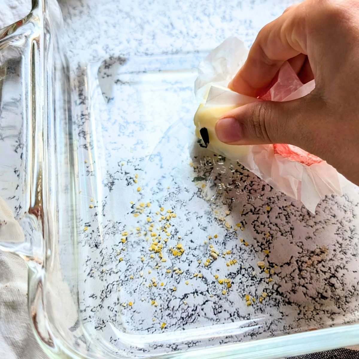 a hand greasing a 8 x 8 baking dish with butter to prep it so the cornbread won't stick.