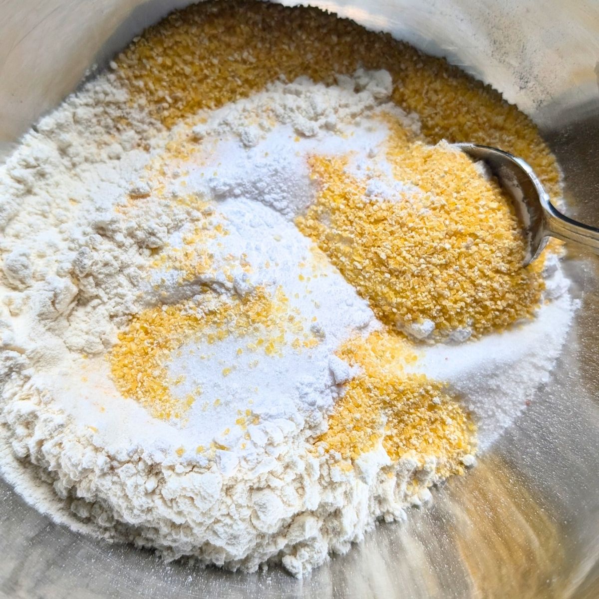 Dry ingredients for cornbread like all putpose flour, cornmeal, sugar, and baking powder, and a little salt mixed with a spoon.