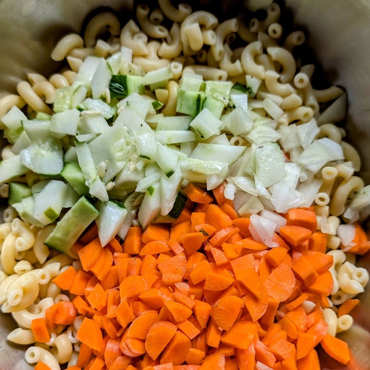 macaroni noodles in a large mixing bowl with chopped cucumbers, carrots, and white onion.