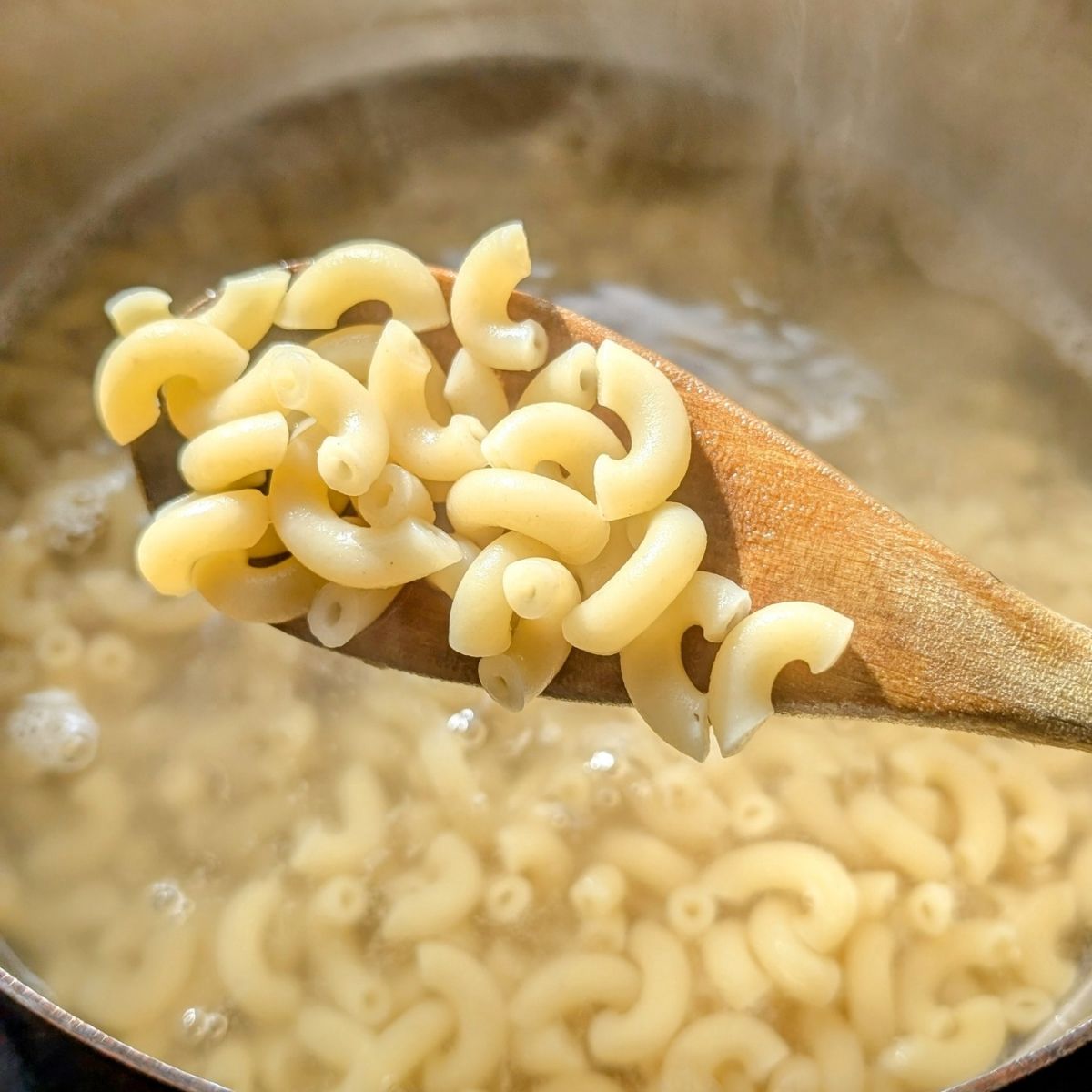 cooked macaroni noodles in a pot being stirred with a wooden spoon to make macaroni salad with cucumbers and dill.