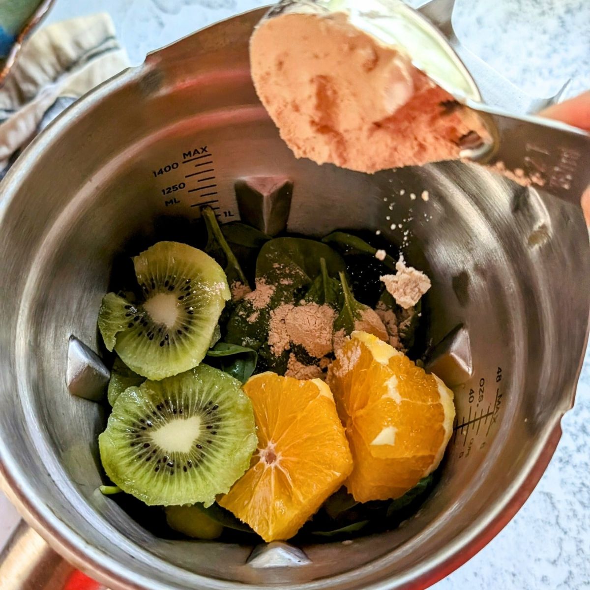 a spoonful of protein powder being added to a stainless steel blender with kiwi and fruit.