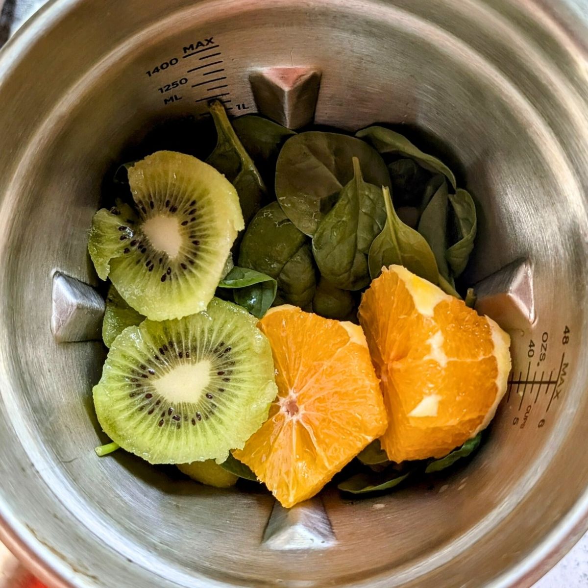 a blender with kiwi oranges spinach banana ready to make a dairy free smoothie in a stainless steel blender.