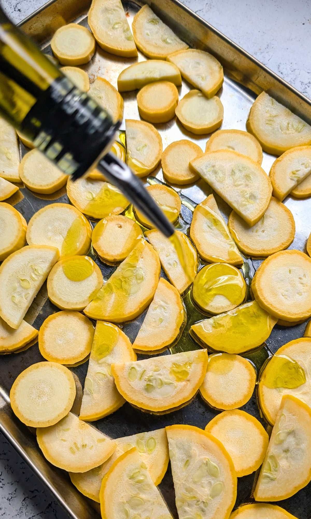 olive oil drizzling over crookneck squash on a sheet pan waiting to be baked.