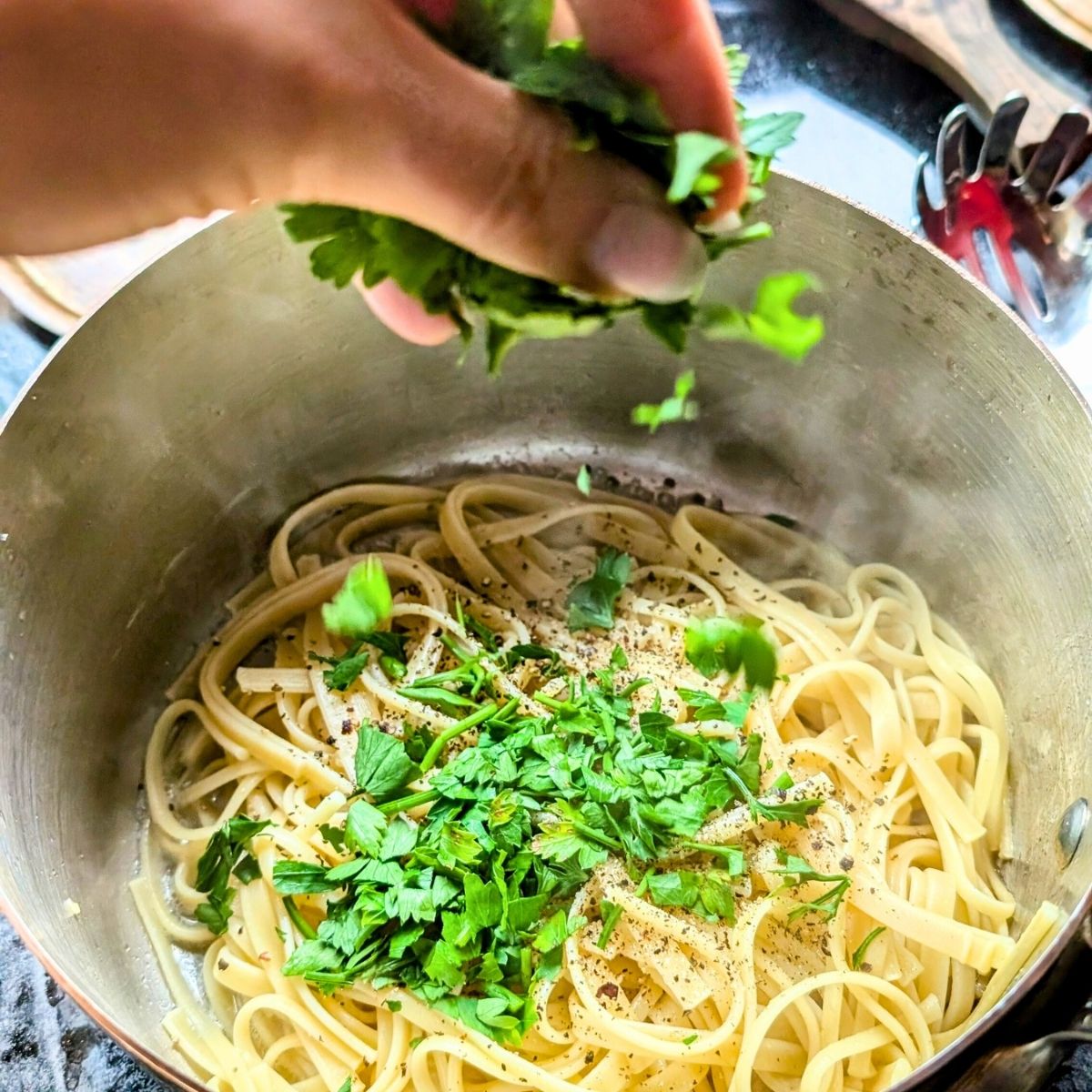 a hand adding fresh chopped parsley to a large pot with cooked noodles.