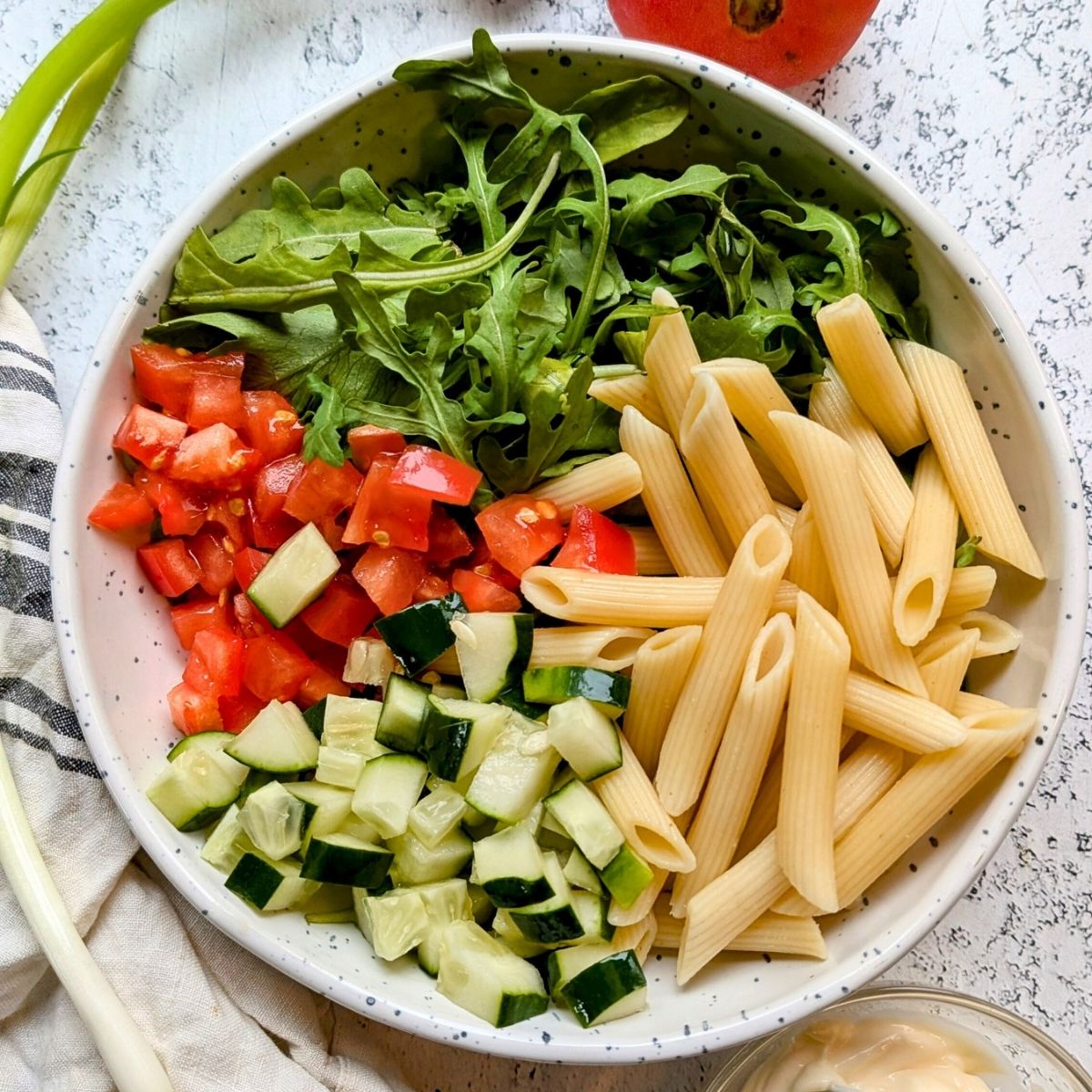 cooked penne pasta in a bowl with fresh arugula, chopped tomatoes, and chopped cucumbers for pasta salad.