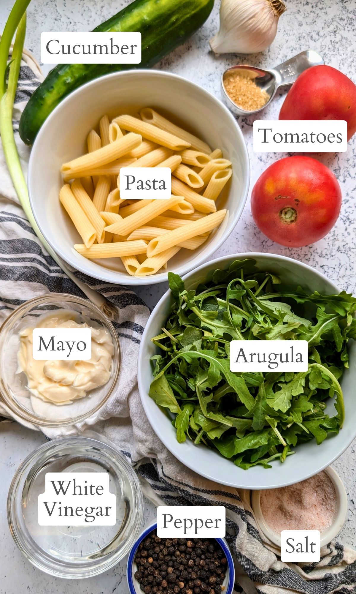 ingredients for arugula pasta salad like penne noodles fresh arugula, tomatoes, cucumber, mayonnaise, and white vinegar for the dressing.