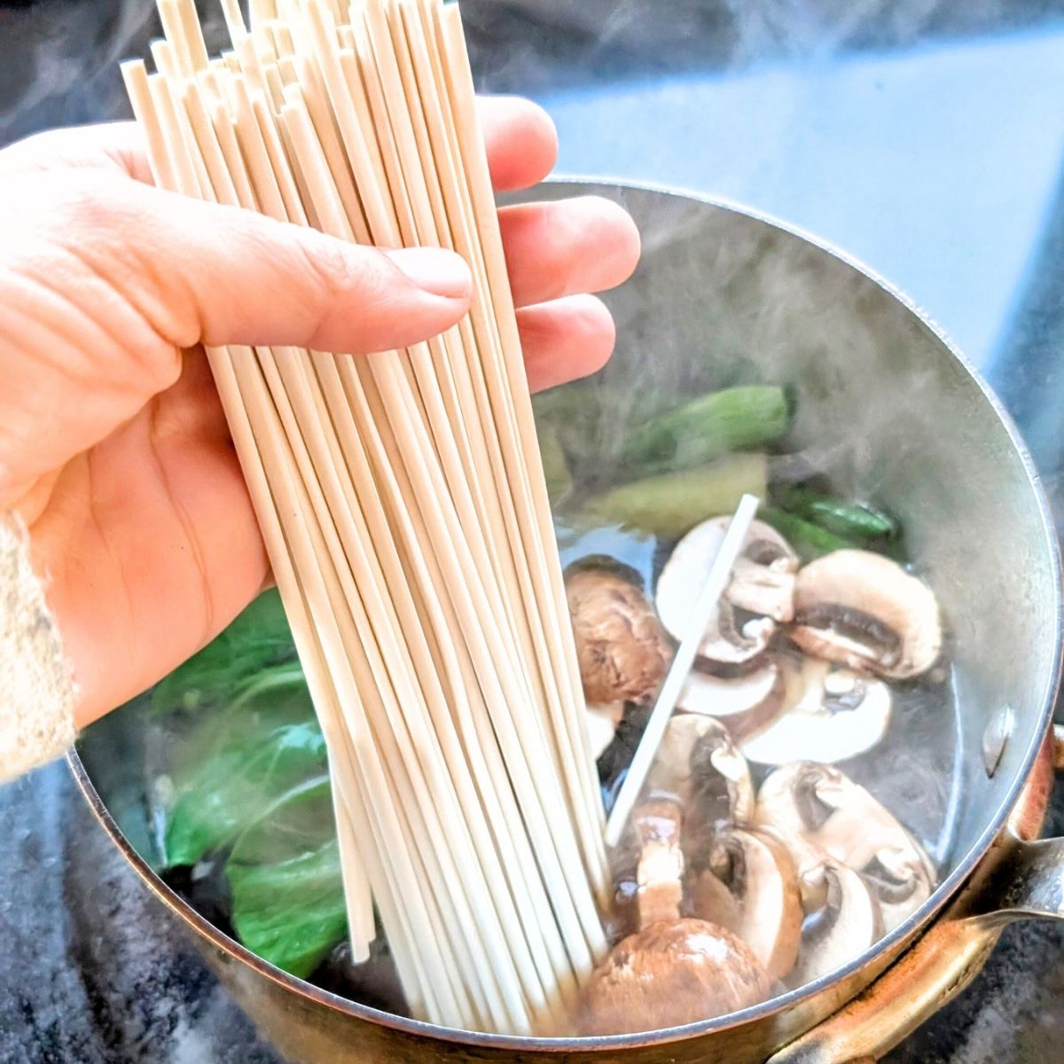 one pot udon soup with dried udon noodles being added to a pot of boiling broth with vegetables.