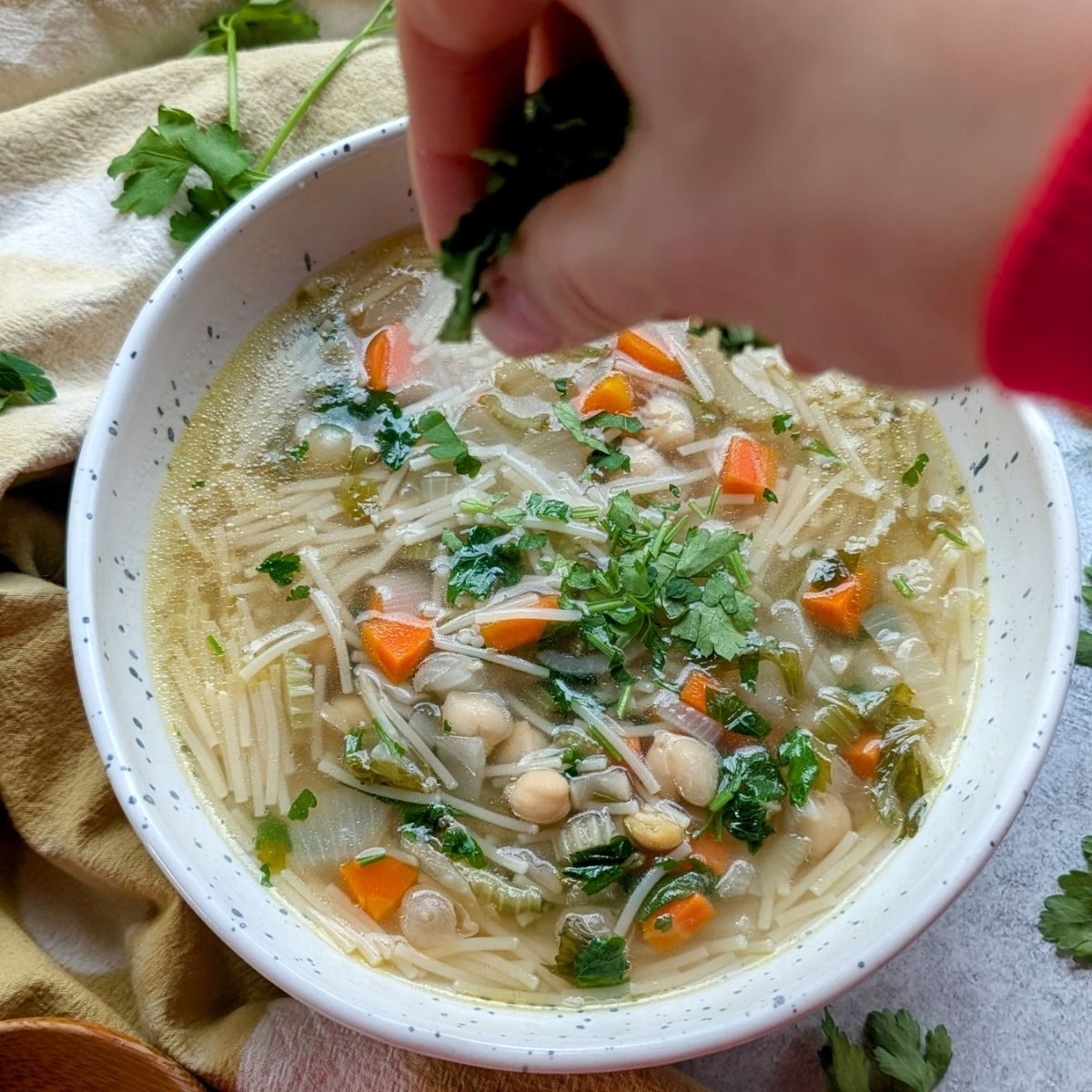 a hand garnishing fresh parsley over soup with noodles and vegetables