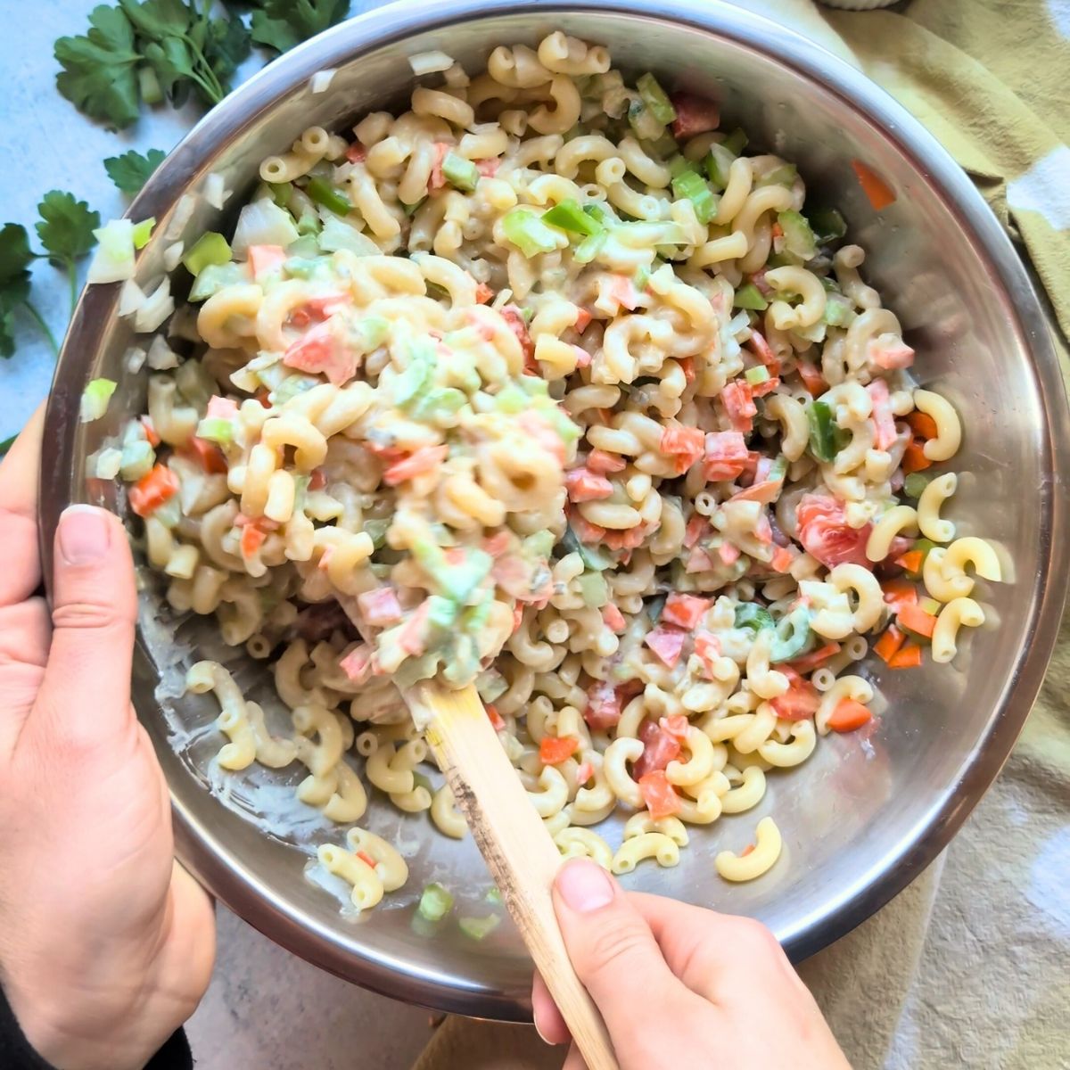 a hand mixing a big bowl of macaroni salad with carrots, onion, celery, and a creamy dressing.