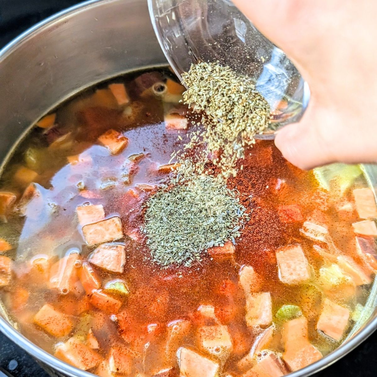 a hand pouring italian seasonings into a pot of split pea soup with sweet potatoes