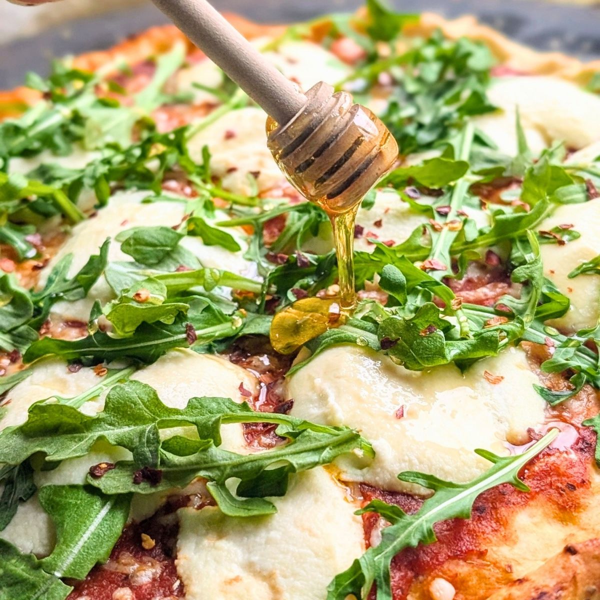 a honey dipper drizzling hot honey over pizza with chili flakes and ricotta cheese.