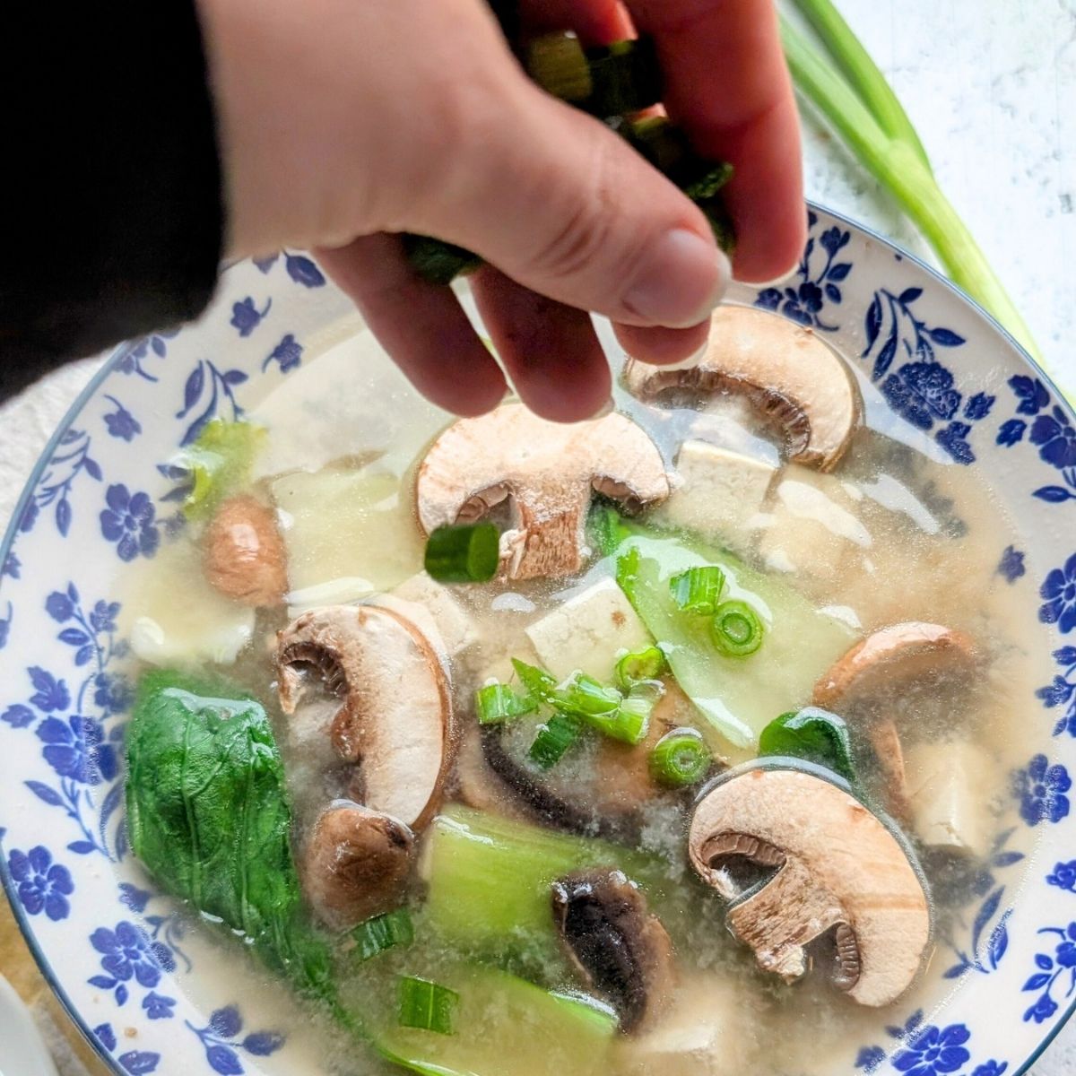 a hand garnishing miso soup with green onions and sliced shiitake mushrooms in miso soup.