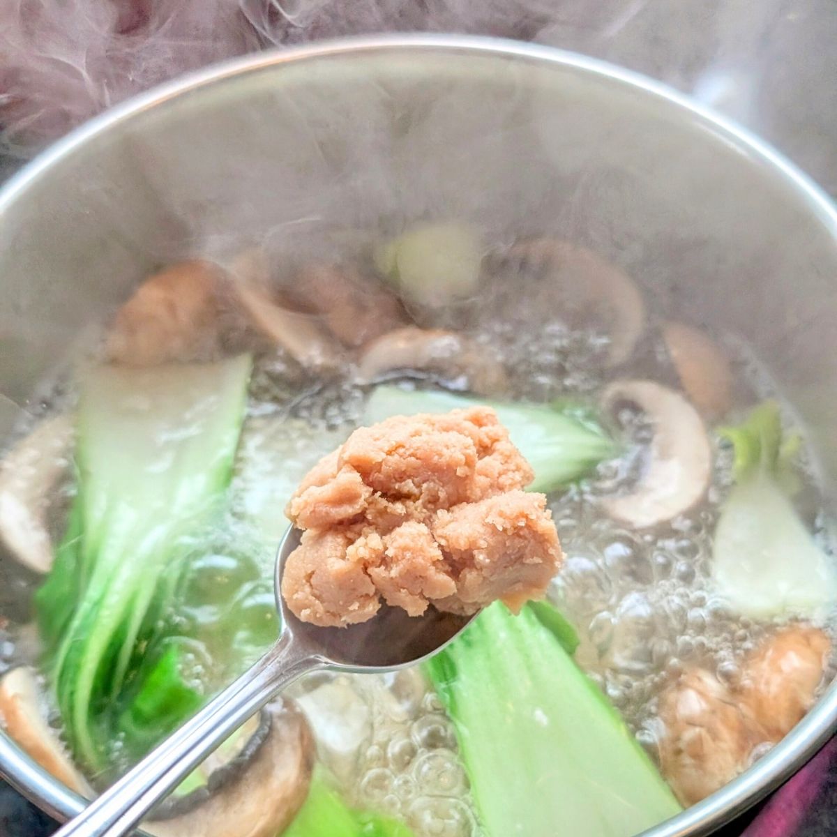 a spoon stirring miso paste into mushroom soup with bok choy for a hearty vegan and vegetarian mushroom soup.