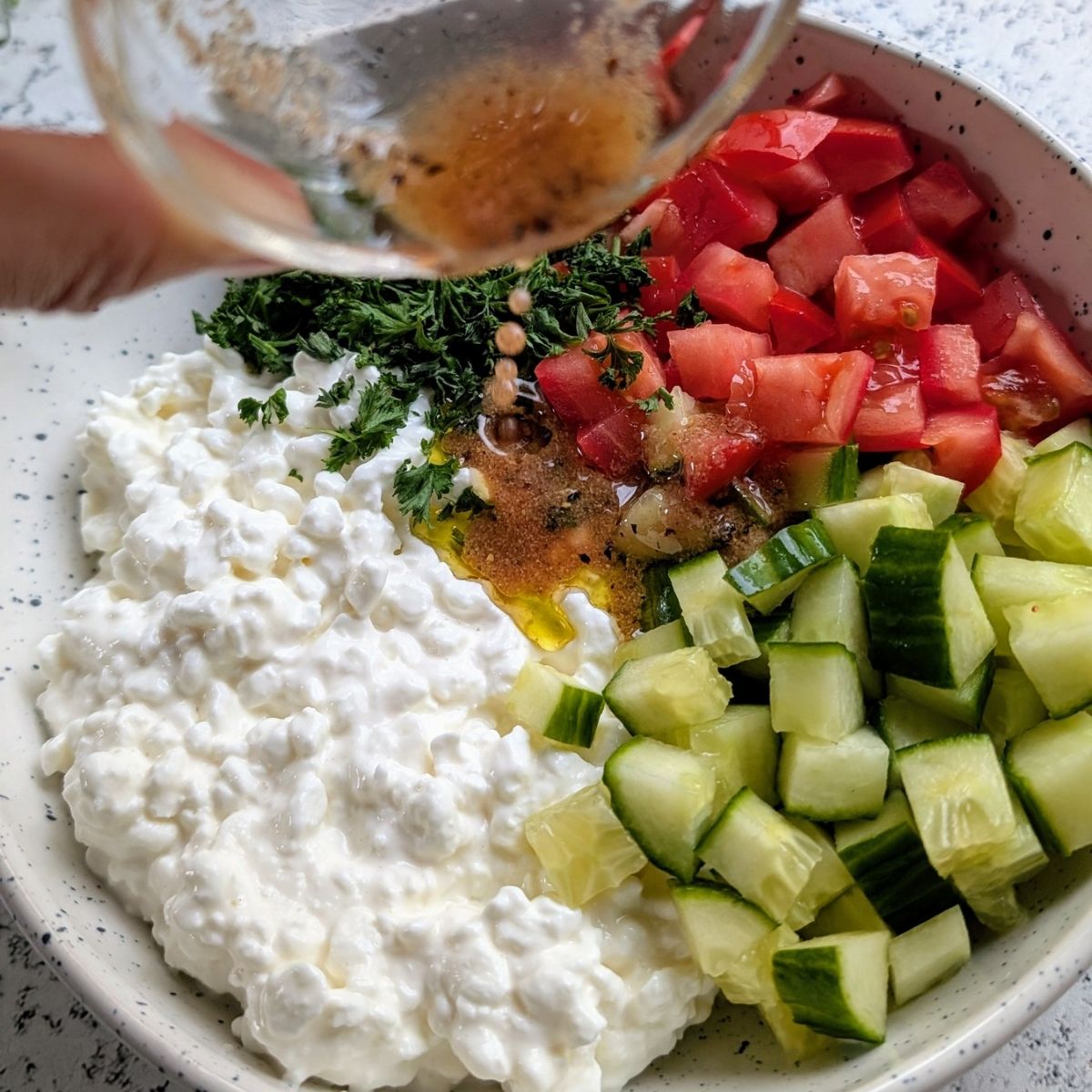 a hand pouring vinaigrette over cottage cheese salad with cucumbers and tomatoes.