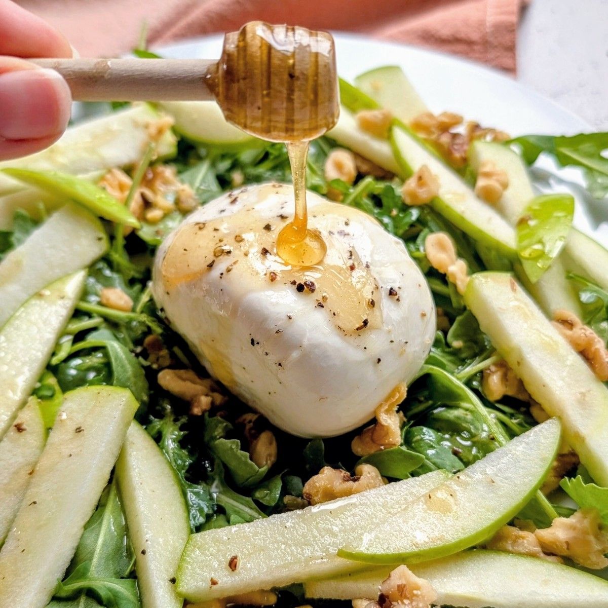 a honey dipper drizzling honey over burrata cheese with apples and greens.