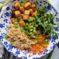 a bowl of sesame tofu with brown rice shredded carrots and arugula with green onions